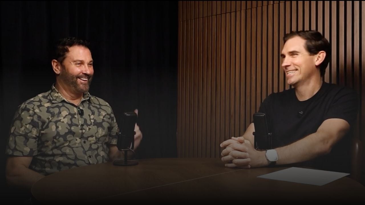 Two men talk across a table with microphones in a podcast-style interview.