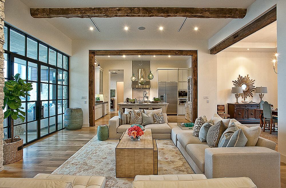 Open-plan living room and kitchen with pale sofas, exposed wood beams, and tall black-framed windows.