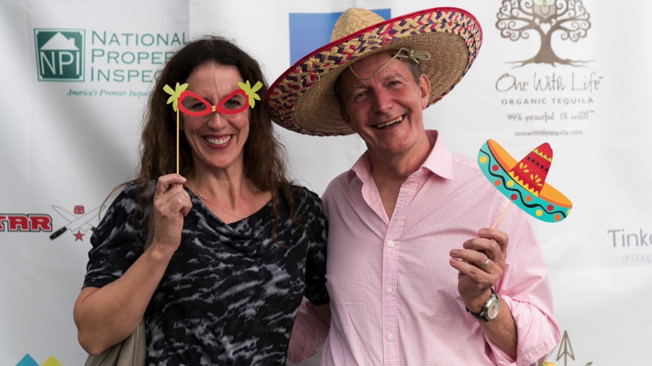 Two adults pose at an event; one wears novelty glasses, and the other wears a colorful sombrero.