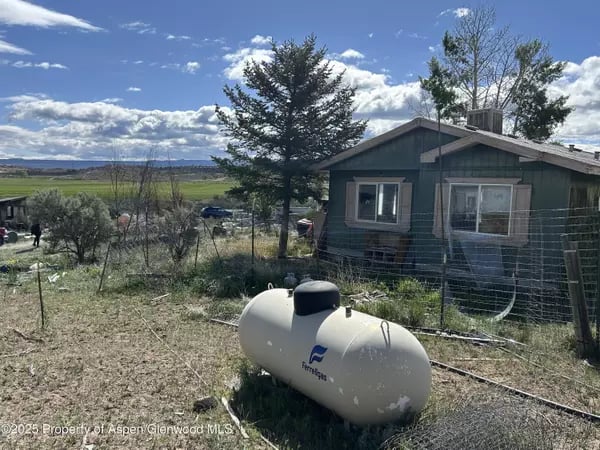 Rural house behind a wire fence, with a white propane tank in the foreground and an open valley beyond.