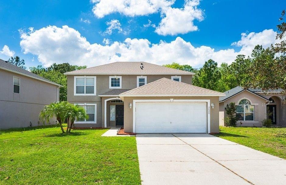 Two-story beige home with a two-car garage, wide driveway, and green lawn.
