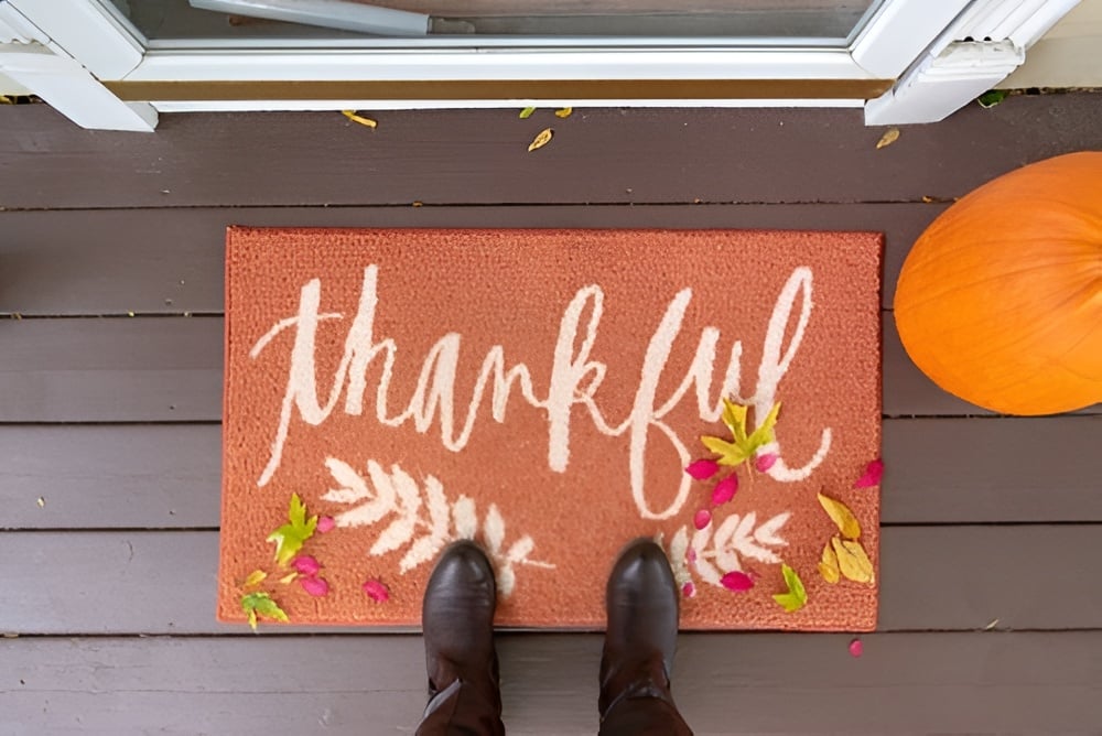 Autumn doorstep with a “thankful” mat, pumpkin, fallen leaves, and dark boots.