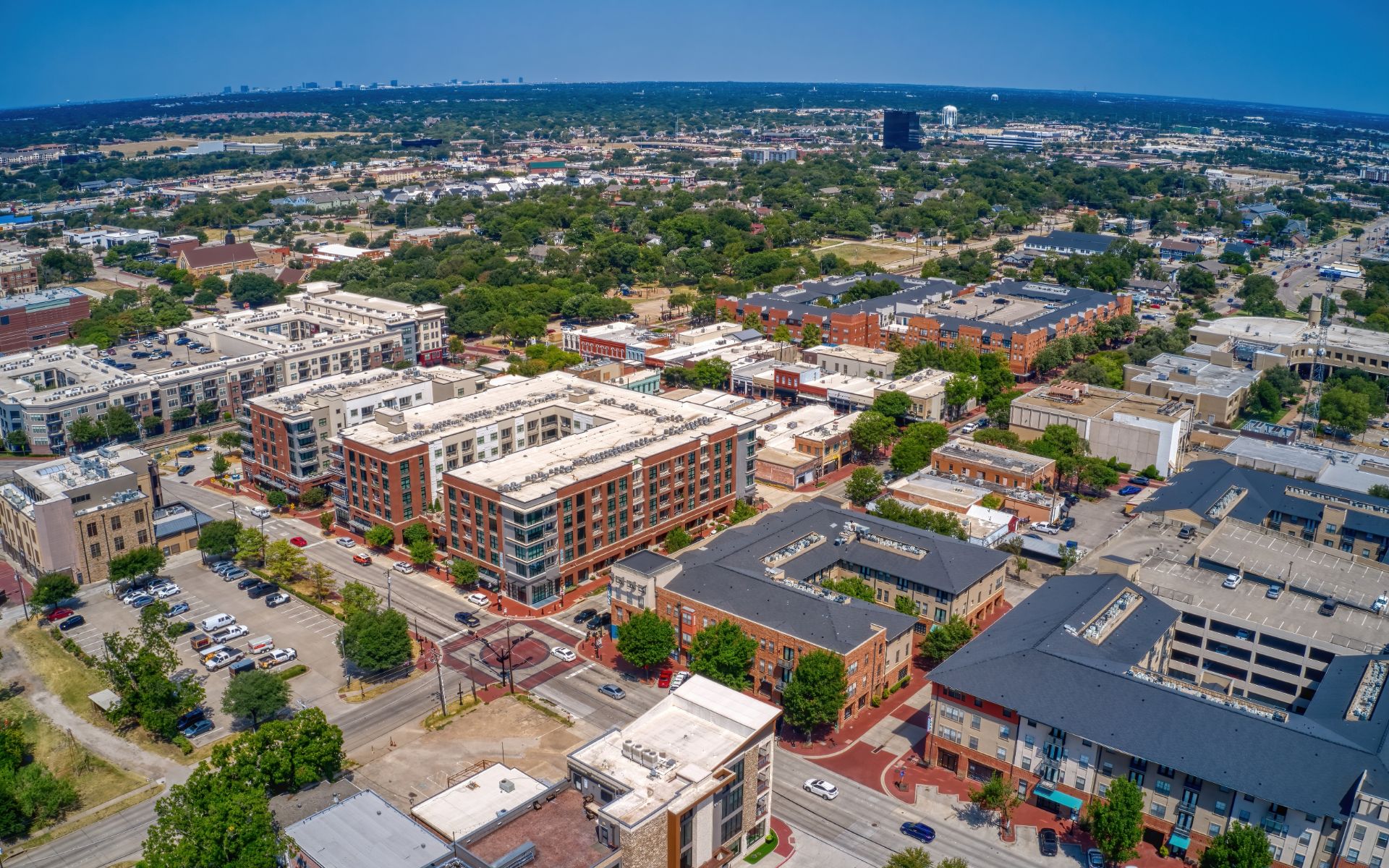 Aerial view of downtown Plano with mid-rise buildings, tree-lined streets, and neighborhoods stretching into the distance.