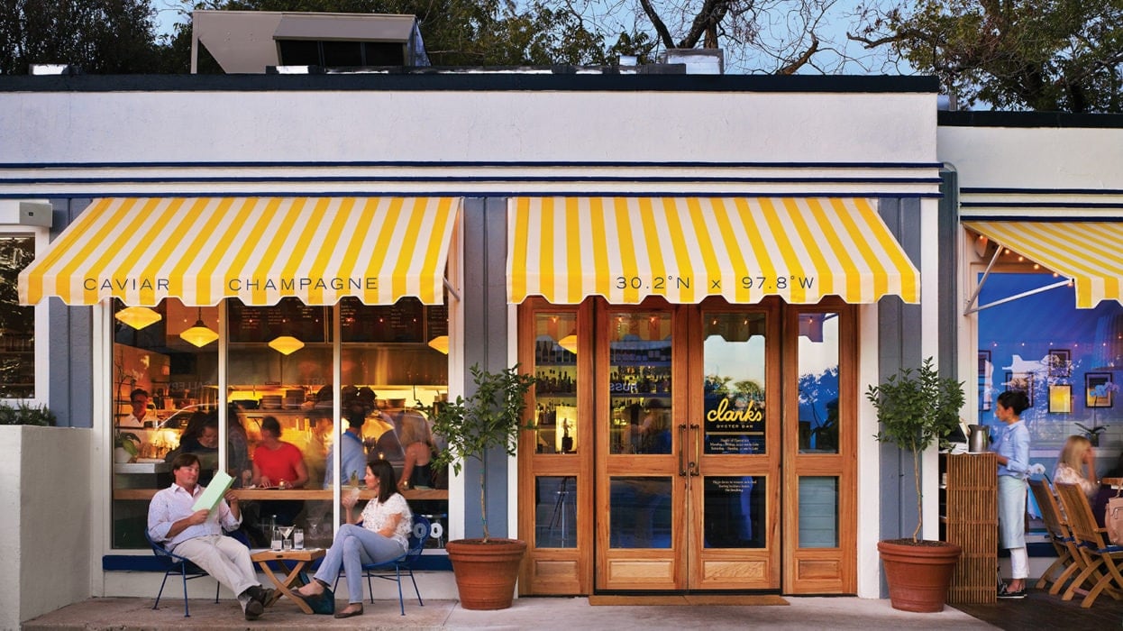 Restaurant storefront with yellow-striped awnings, wooden doors, and diners seated outside.
