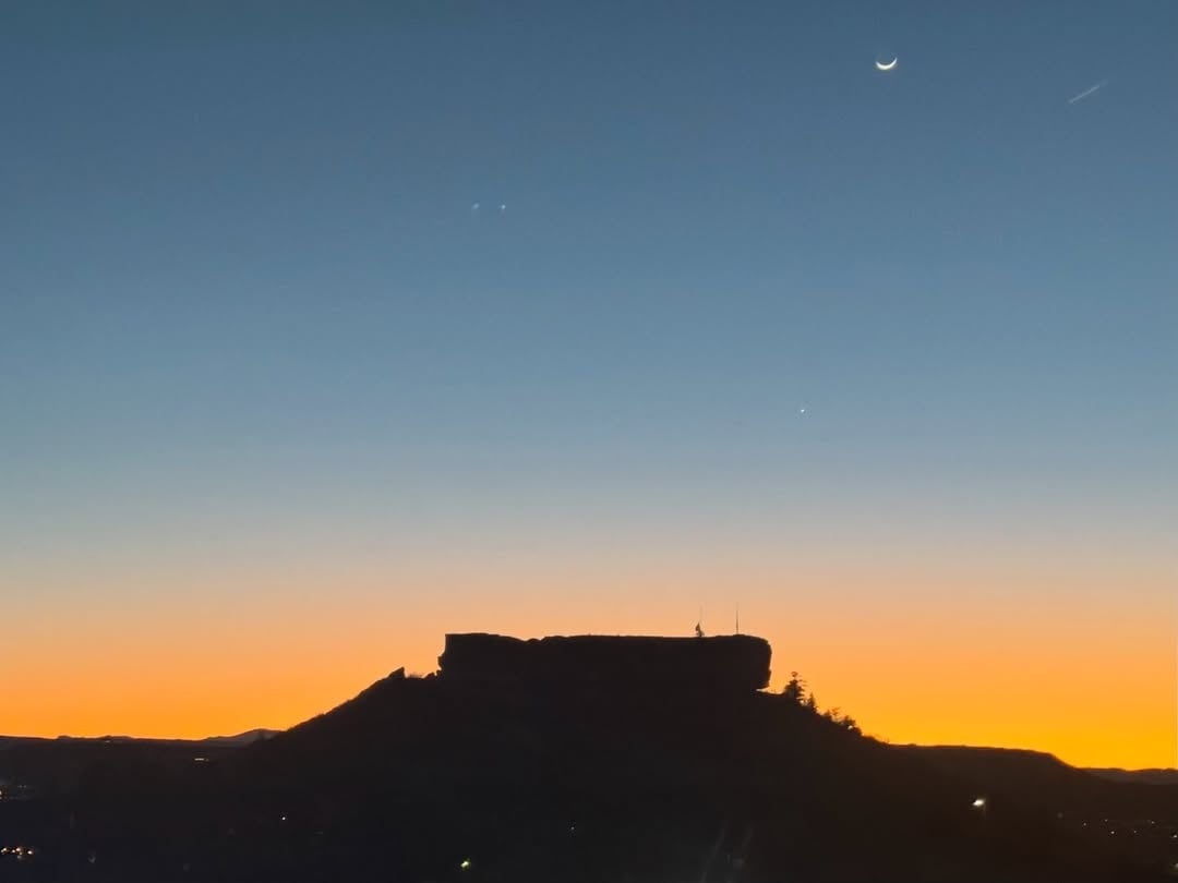 Dramatic sunset silhouette of Castle Rock formation with town lights emerging below and foothill panorama.