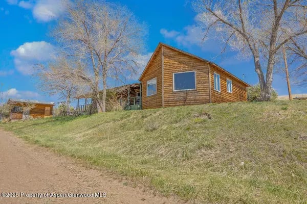 Wood-sided cabin on a grassy slope beside a dirt drive, with a small outbuilding nearby.