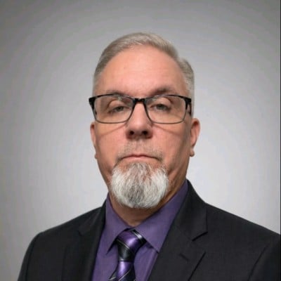 Kevin Pattison wearing glasses, a dark suit, and a purple shirt and tie.
