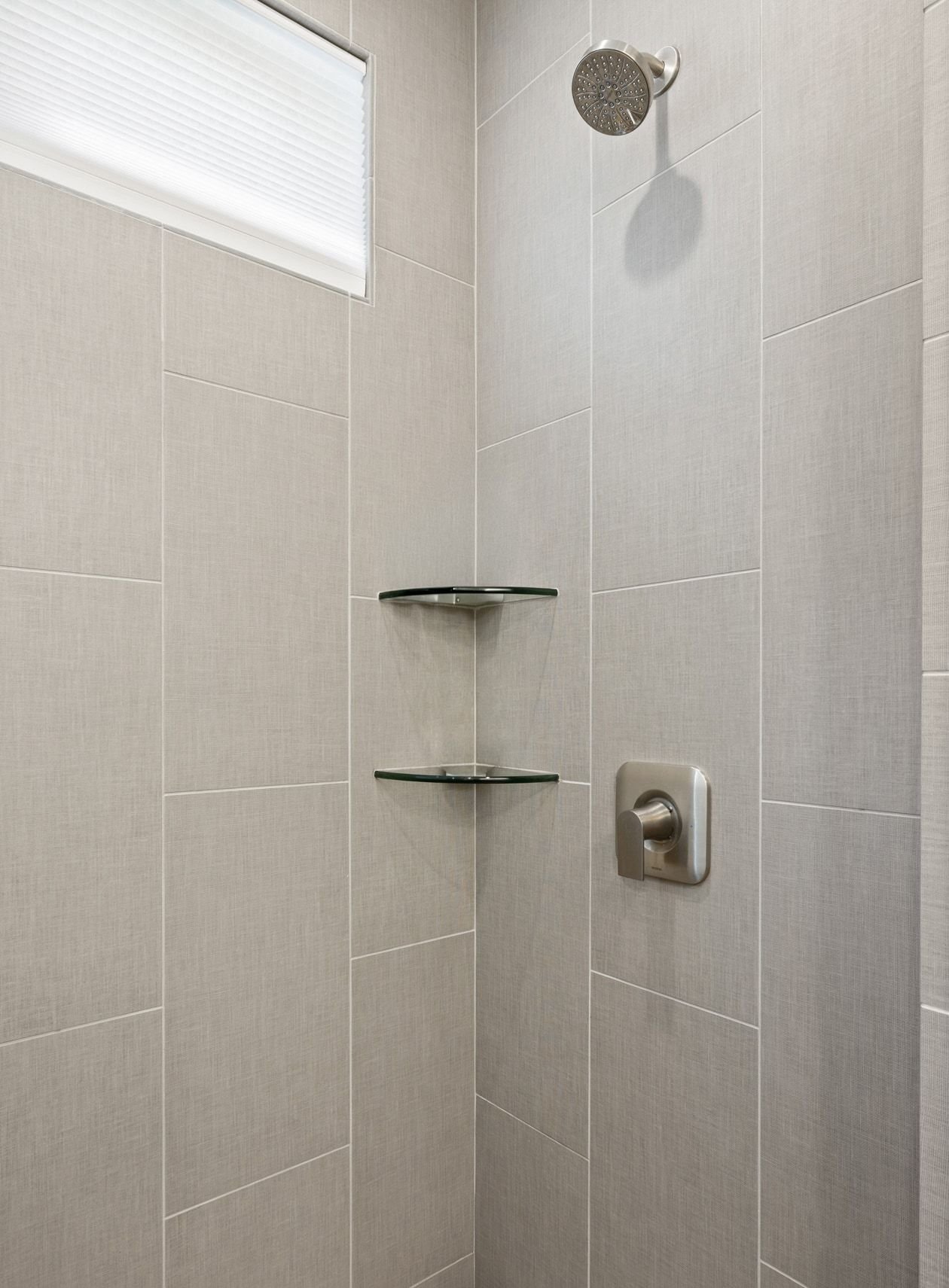 Tiled shower with a rainfall showerhead, glass corner shelves, and a shaded window.