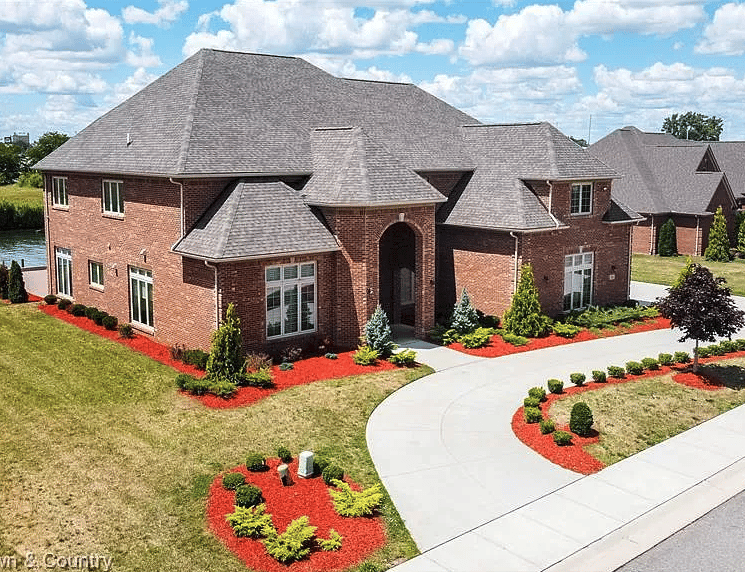 Large red-brick home with a gray roof, arched entry, landscaped gardens, and curved driveway.