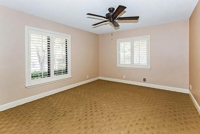 Empty bedroom with tan patterned carpet, shuttered windows, and a dark ceiling fan.