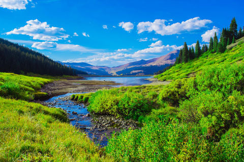 Mountain stream winds through green vegetation toward a lake beneath distant peaks.