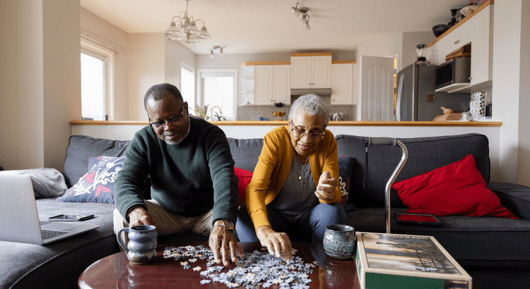 Elderly couple sitting on a couch, working on a jigsaw puzzle on a coffee table. They are relaxed and smiling, surrounded by cozy home decor.