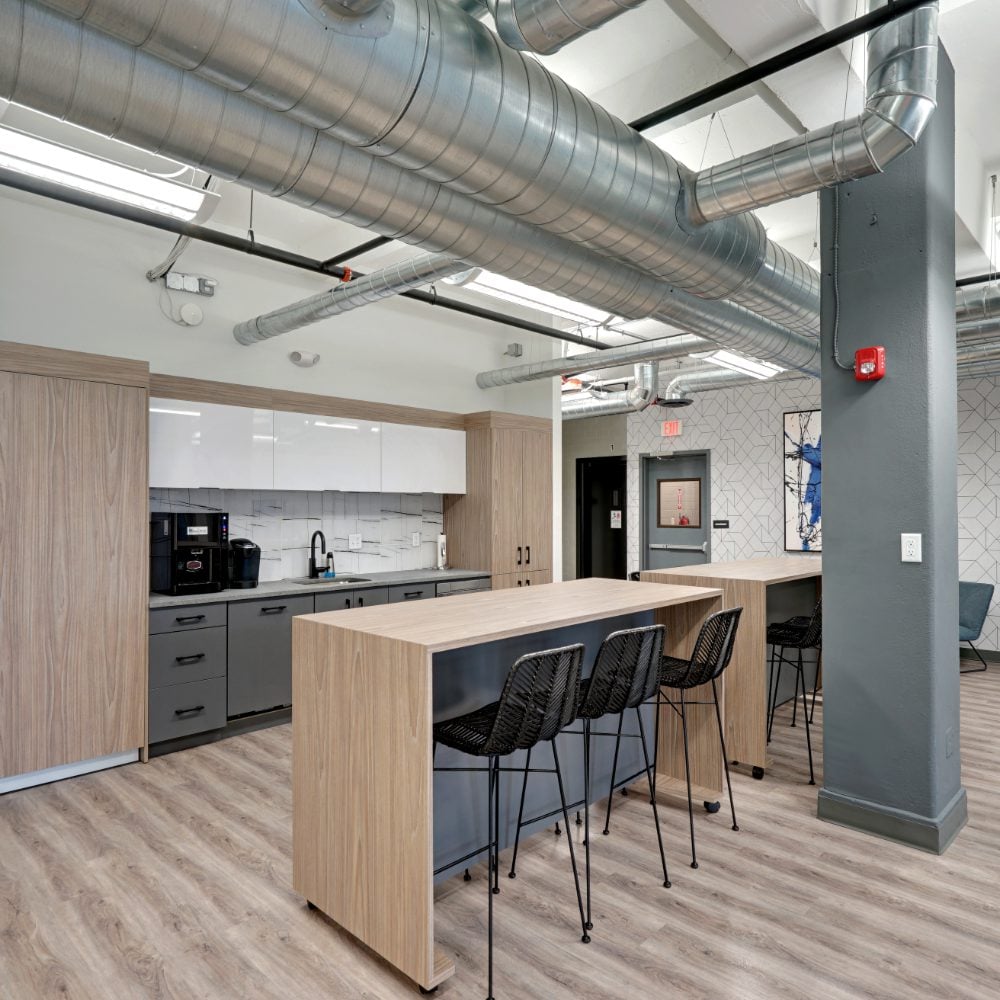 Shared kitchen with a large wood-finish island, tall black chairs, and exposed ceiling ducts.