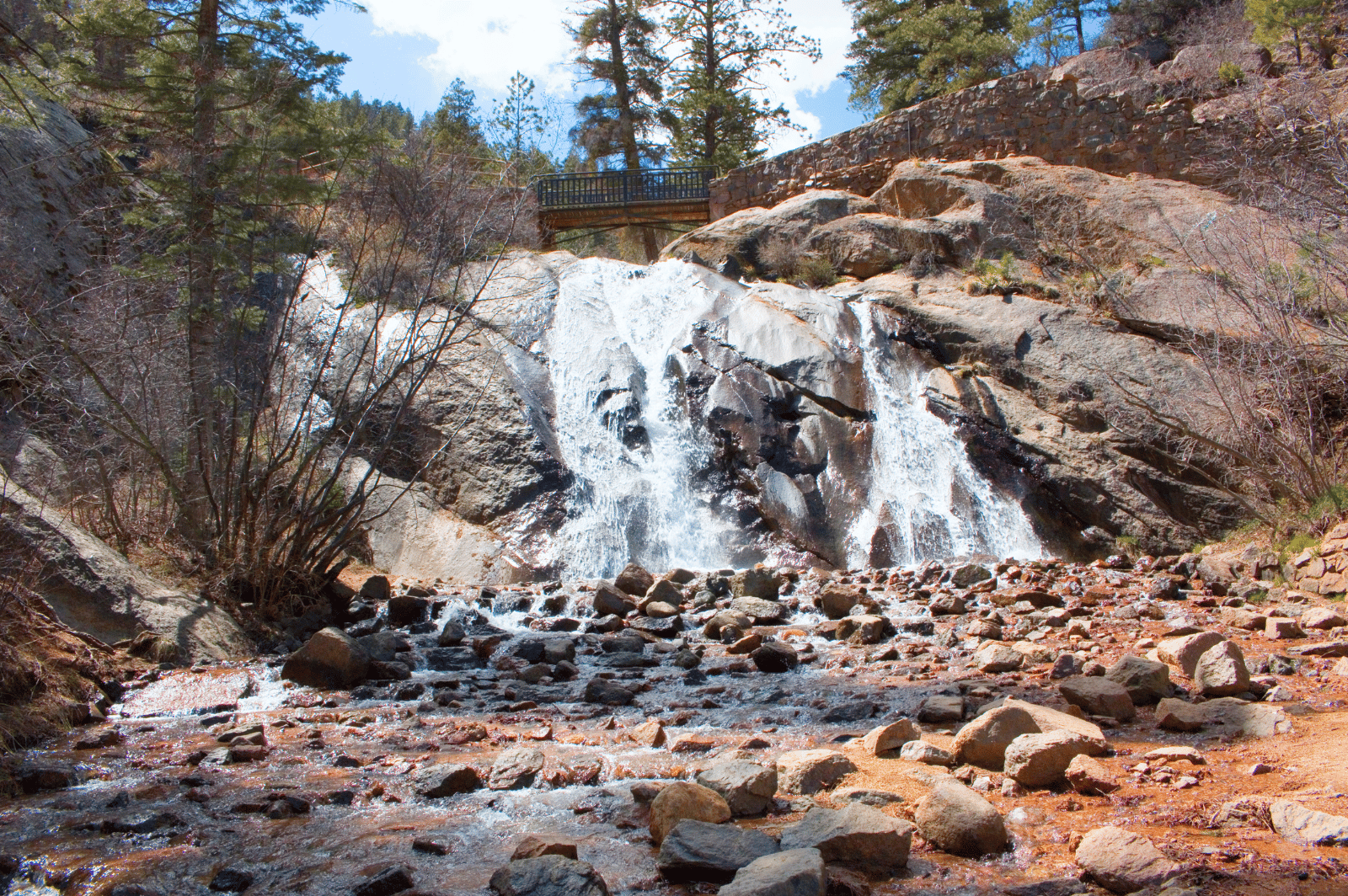 Waterfall over rocky ledges beneath a pedestrian bridge, flowing into a boulder-strewn stream.
