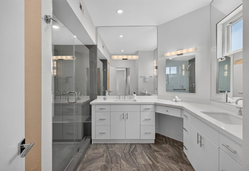 Modern bathroom with a glass shower, long white vanity, multiple sinks, and dark patterned tile flooring.
