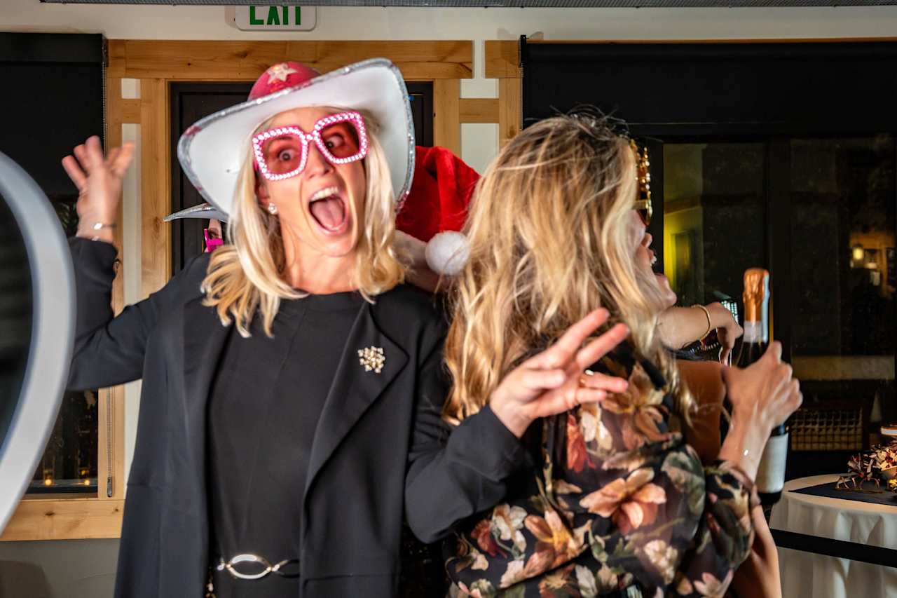 Woman in a white cowboy hat and oversized pink glasses celebrates beside a woman in a floral dress.