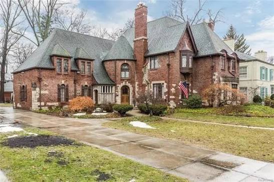 Red-brick Tudor Revival house with steep slate roofs, prominent chimneys, and patches of snow on the lawn.