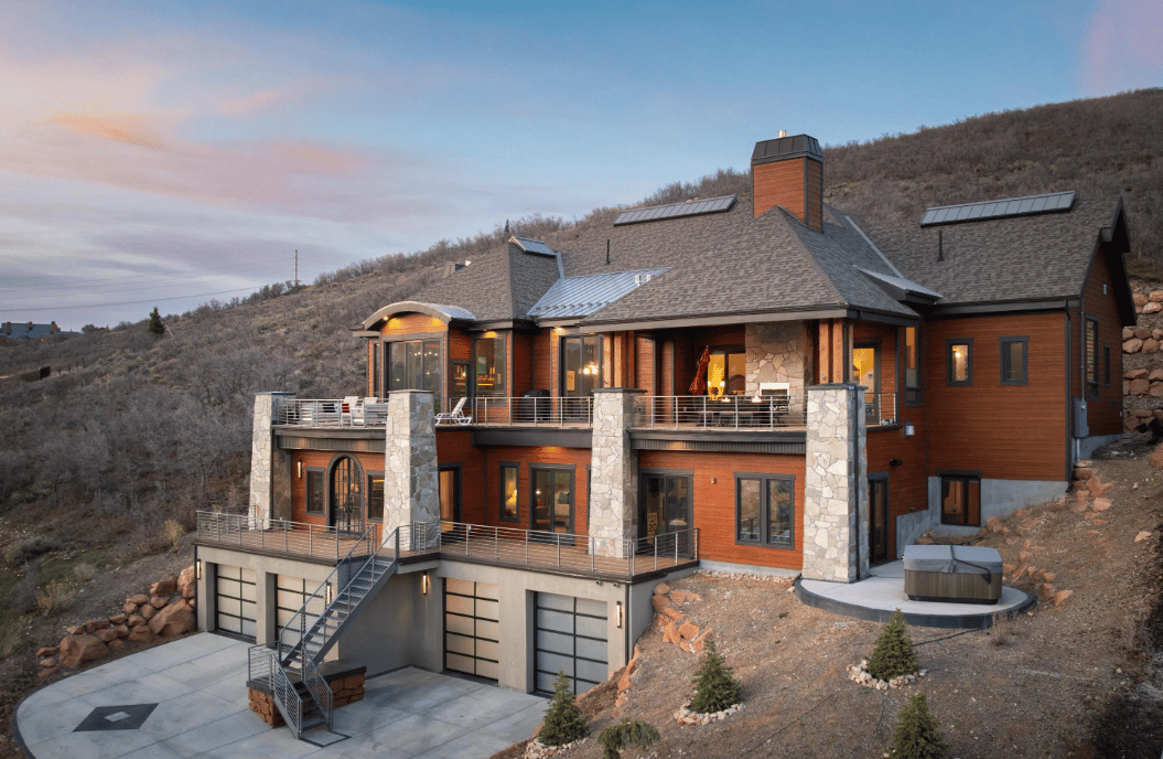 Hillside home with broad decks, stone columns, exterior stairs, and a hot tub at dusk.