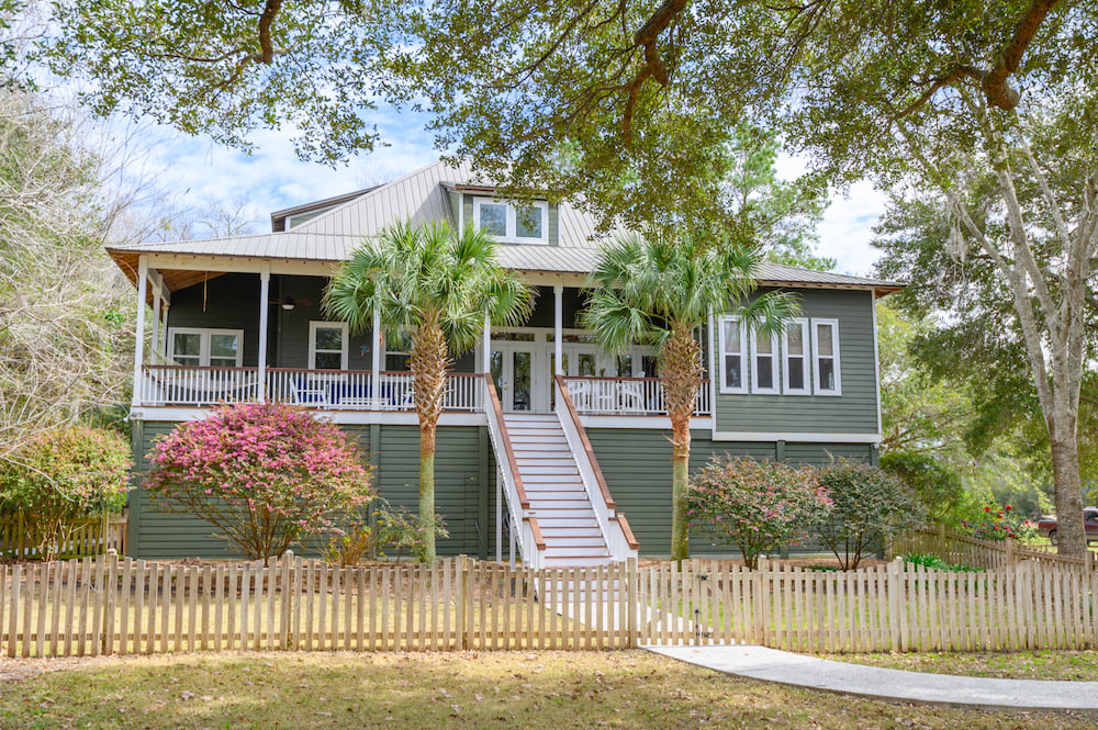 Raised green home with a broad porch, central stairs, palm trees, and white picket fence.