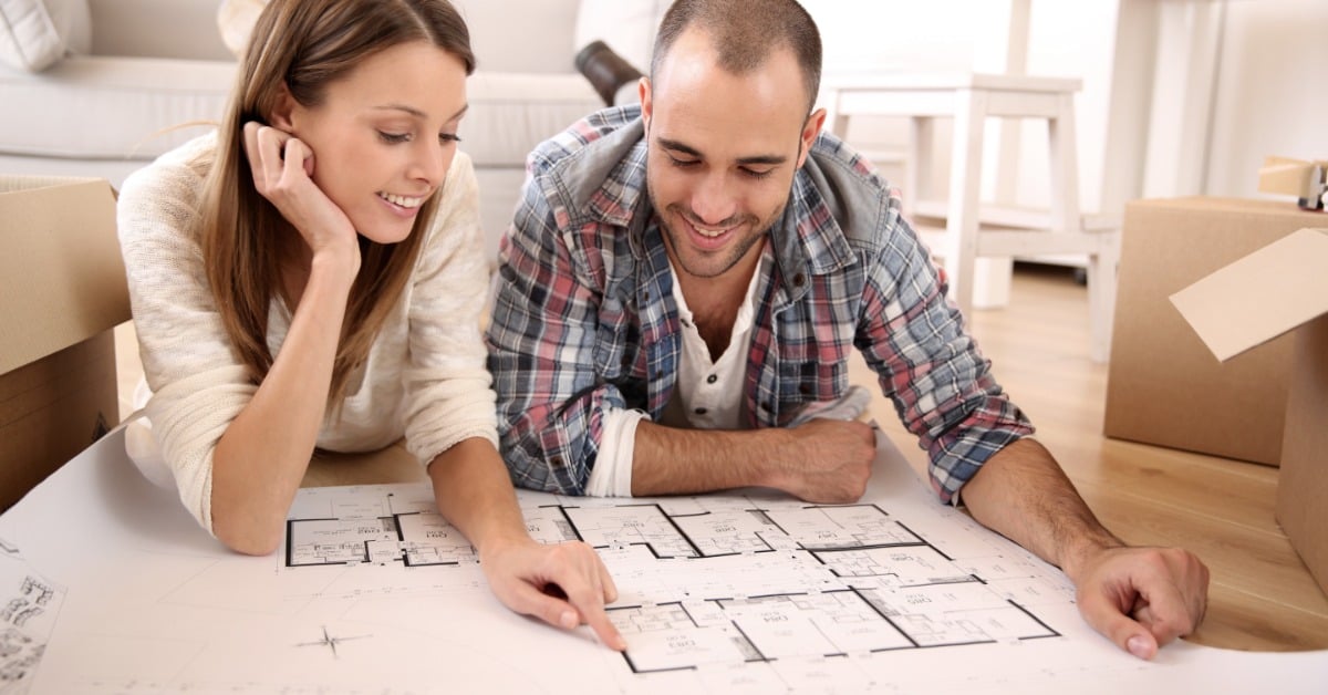 A man and a woman laying stomach-first on the hardwood floor of a home looking at architecture drawings.