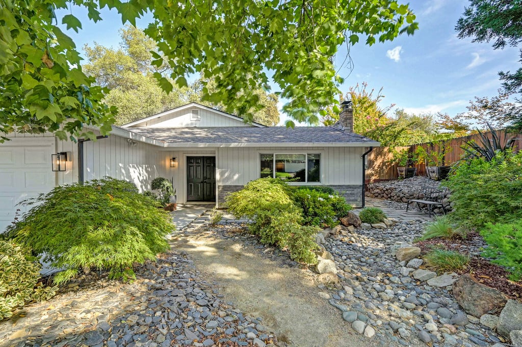 Single-story gray home with a black front door, stone landscaping, and mature trees.