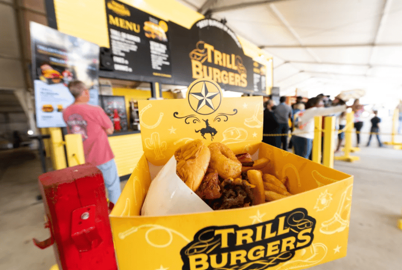 Burger and fries in a yellow Trill Burgers box at a busy rodeo food stand.
