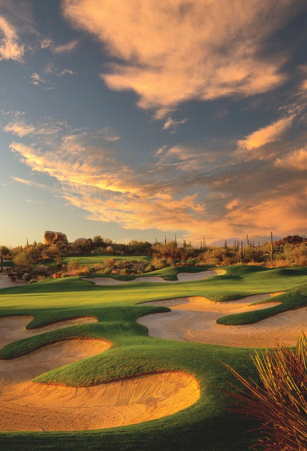 Golf course fairways and sand bunkers beneath dramatic orange sunset clouds.