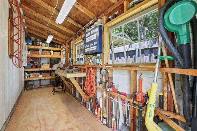 Wooden workshop with a long workbench, stocked shelves, hanging tools, and side windows.