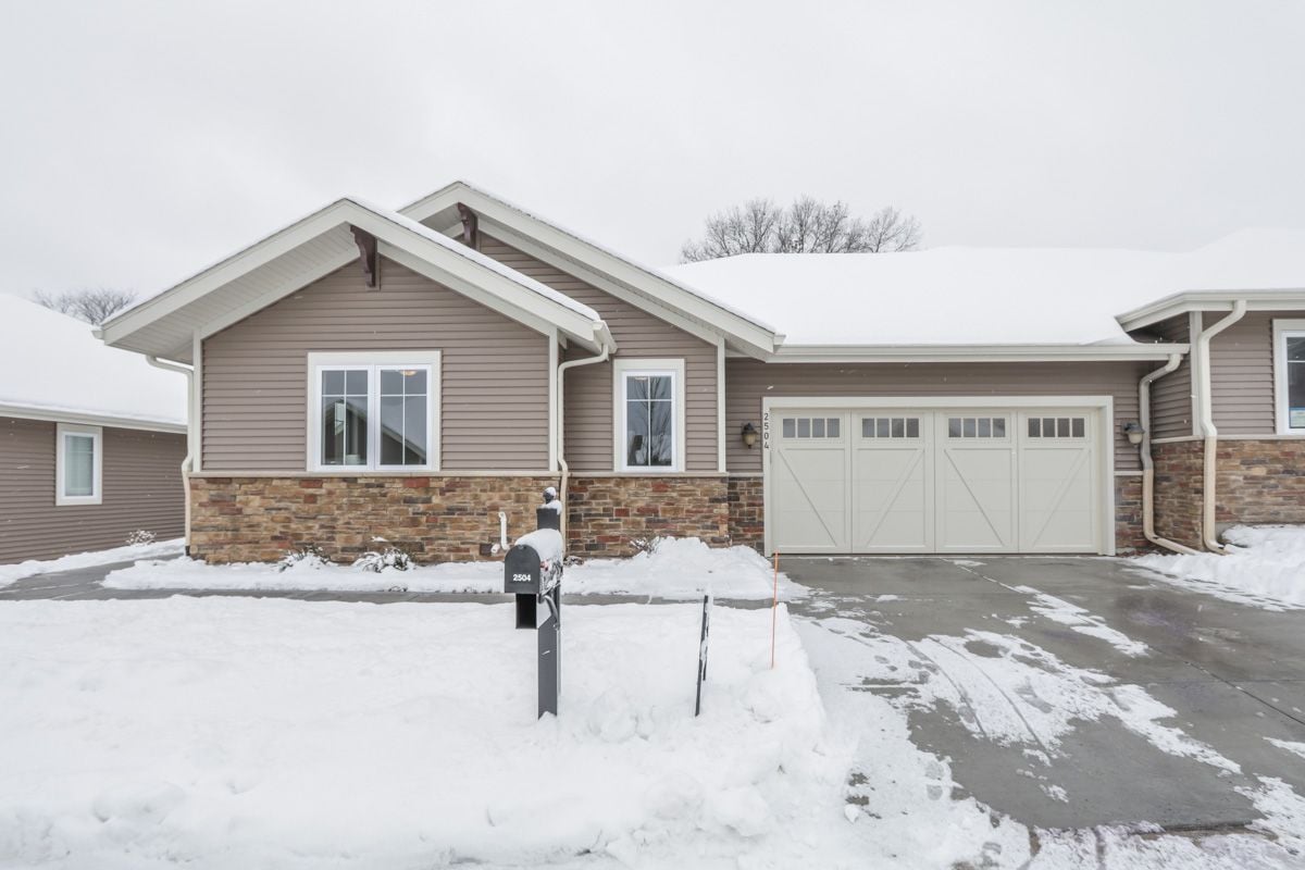 Snow-covered home with taupe siding, stone facade, and a two-car garage.