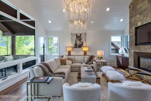 Living room with a cream sectional, stone fireplace, and large windows overlooking greenery.