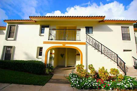Two-story cream residential building with an exterior staircase, arched entrance, and flower beds.