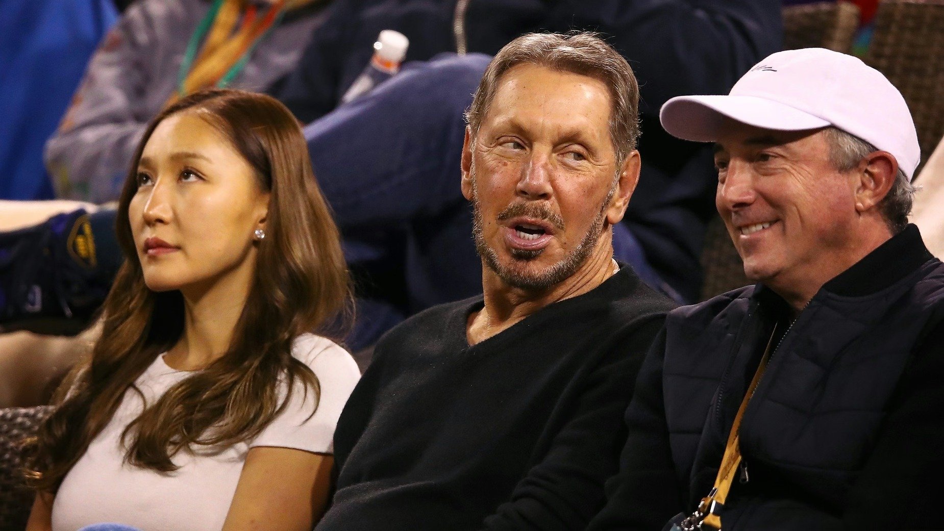 Larry Ellison seated between a woman and a man in a white cap at a sporting event.