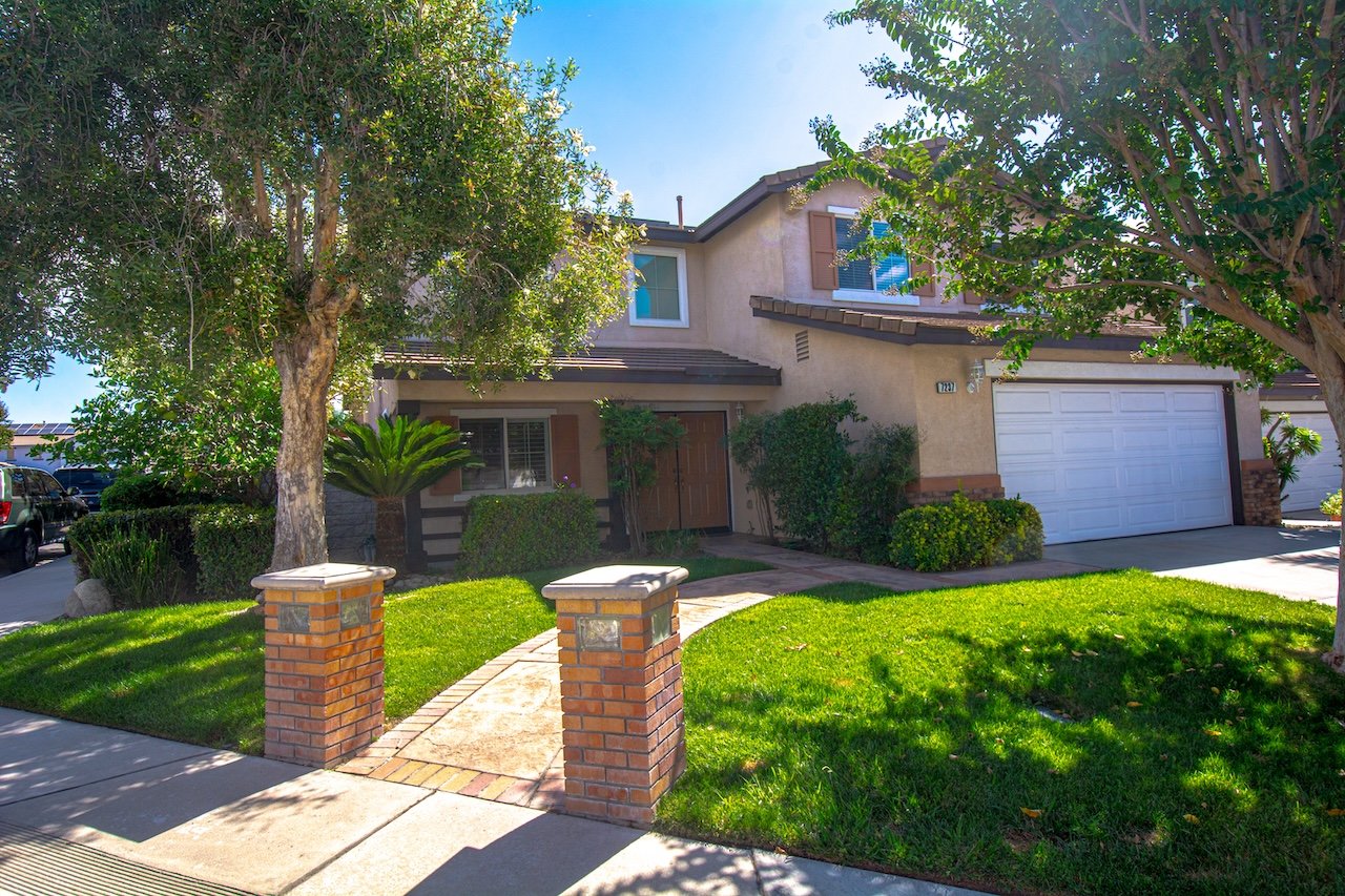 Two-story beige stucco house with a white garage door, mature trees, and a brick-edged walkway.