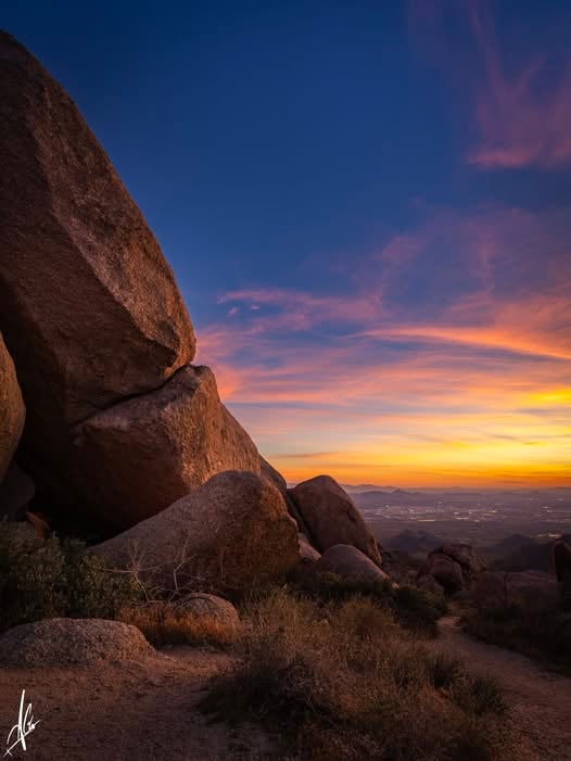 Desert boulders and scrub frame distant mountains beneath a colorful sunset.