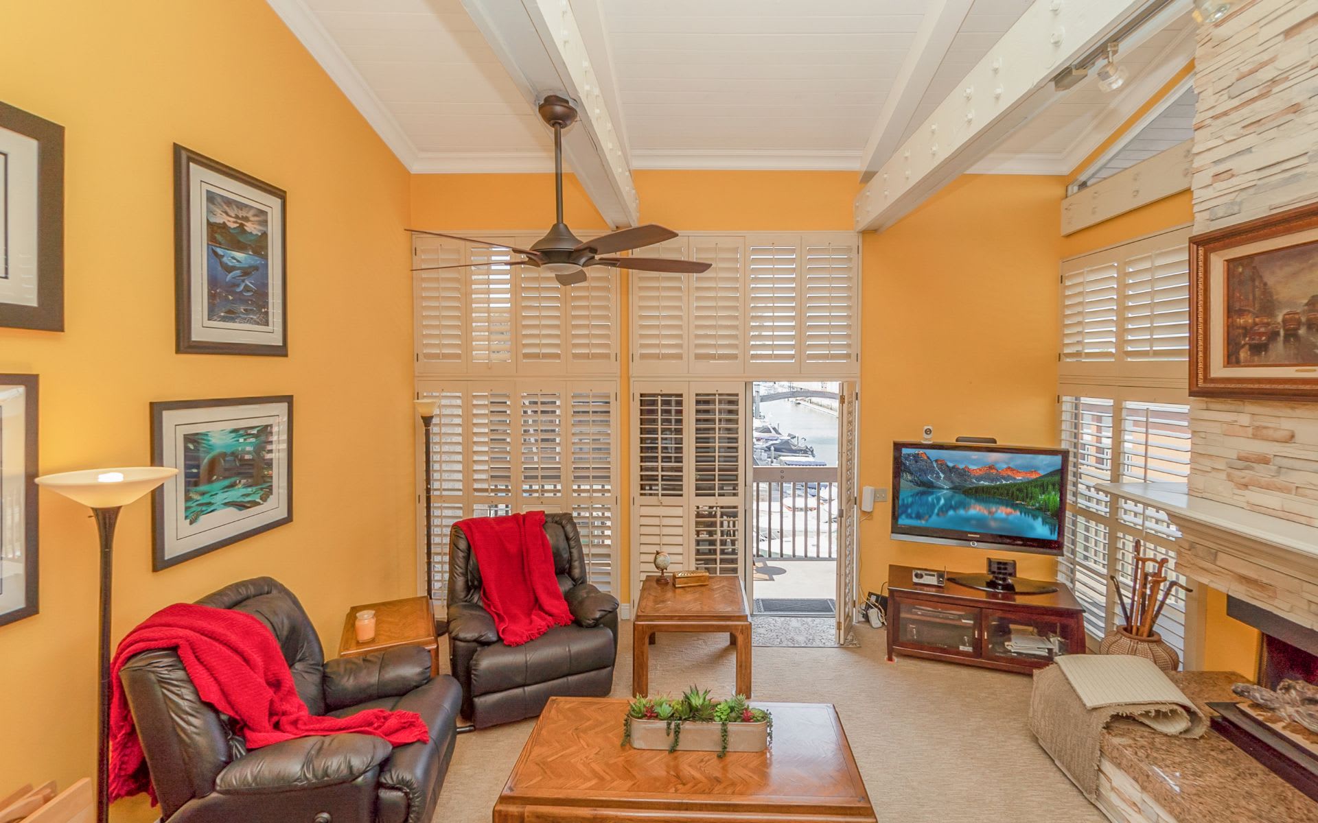 Living room with white ceiling beams, leather recliners, stone fireplace, and marina view through the doors.