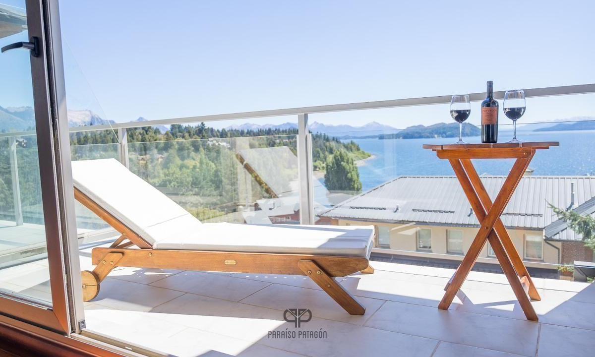 Apartment balcony with a wooden sun lounger overlooking a lake and distant mountains.