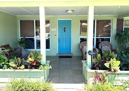 Green house with a bright blue front door, covered porch, wicker chairs, and potted plants.