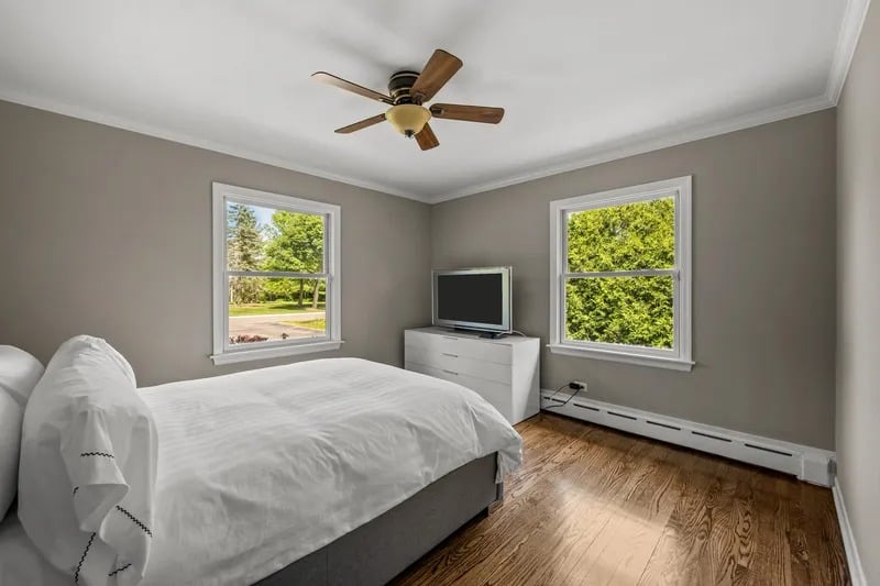 Bedroom with a white bed, hardwood floors, and two windows overlooking green trees.