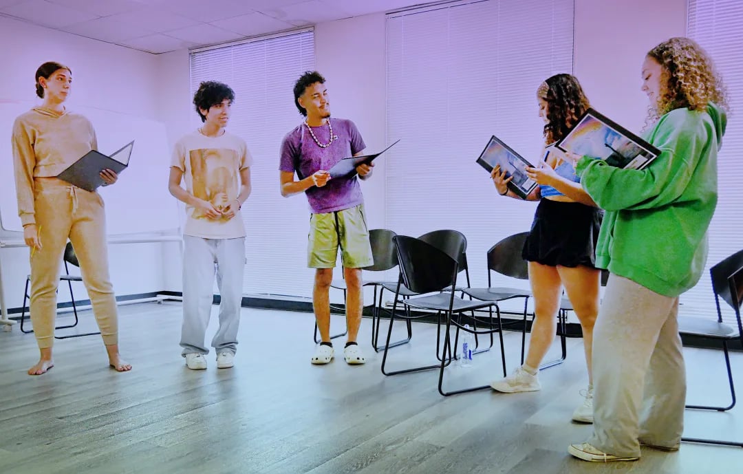 Five young people discuss images on tablets in a bright room with chairs.