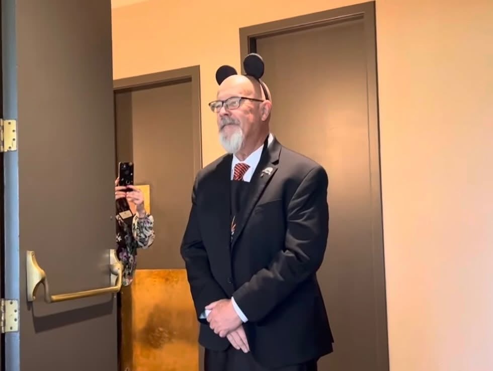 Bearded man in a dark suit and red tie wearing round mouse ears, with someone photographing him behind a door.