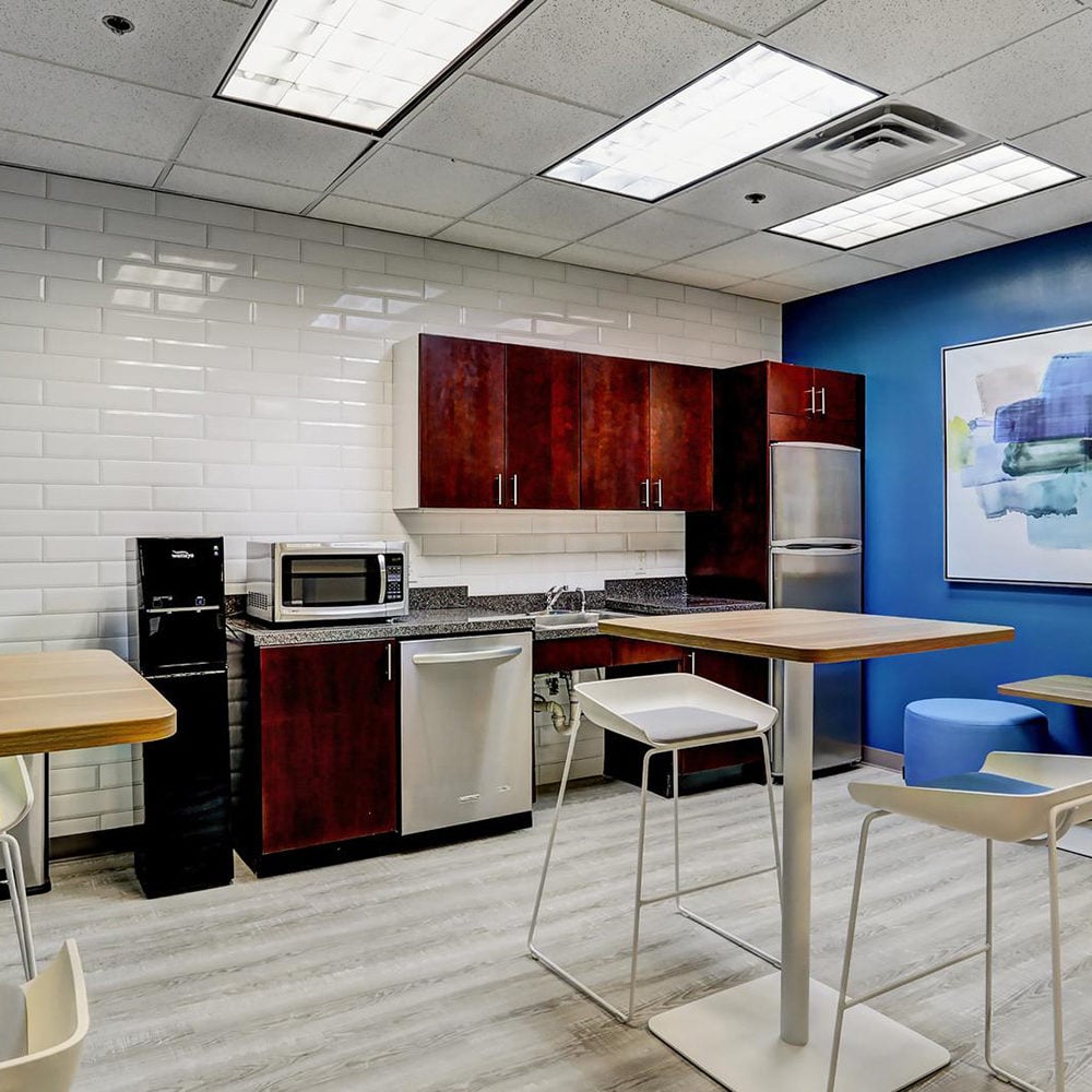 Kitchenette with dark wood cabinets, stainless appliances, and tall tables in a blue-accented break area.