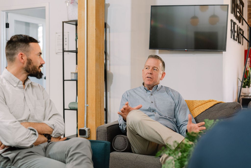 Two men talk in an office lounge, one gesturing from a gray sofa while the other listens.