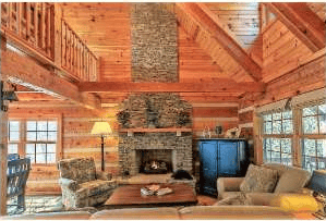 Log cabin living room with a stone fireplace, exposed timber beams, and large windows.