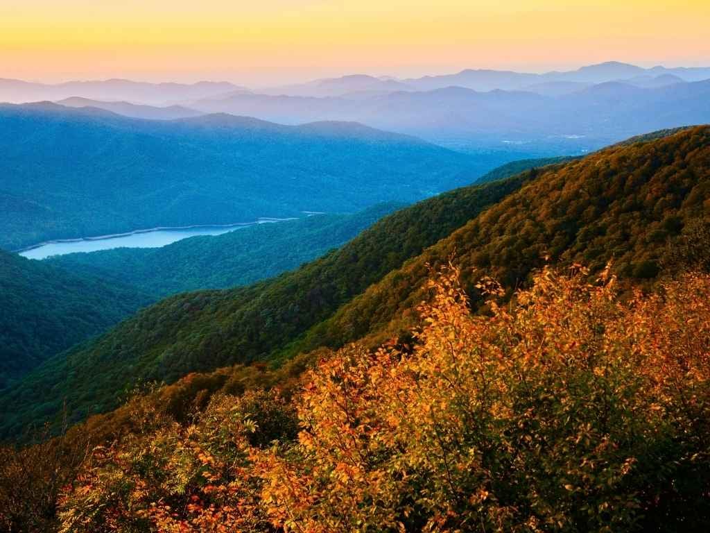 Sunset over layered mountain ridges and a valley lake, with fall foliage in the foreground