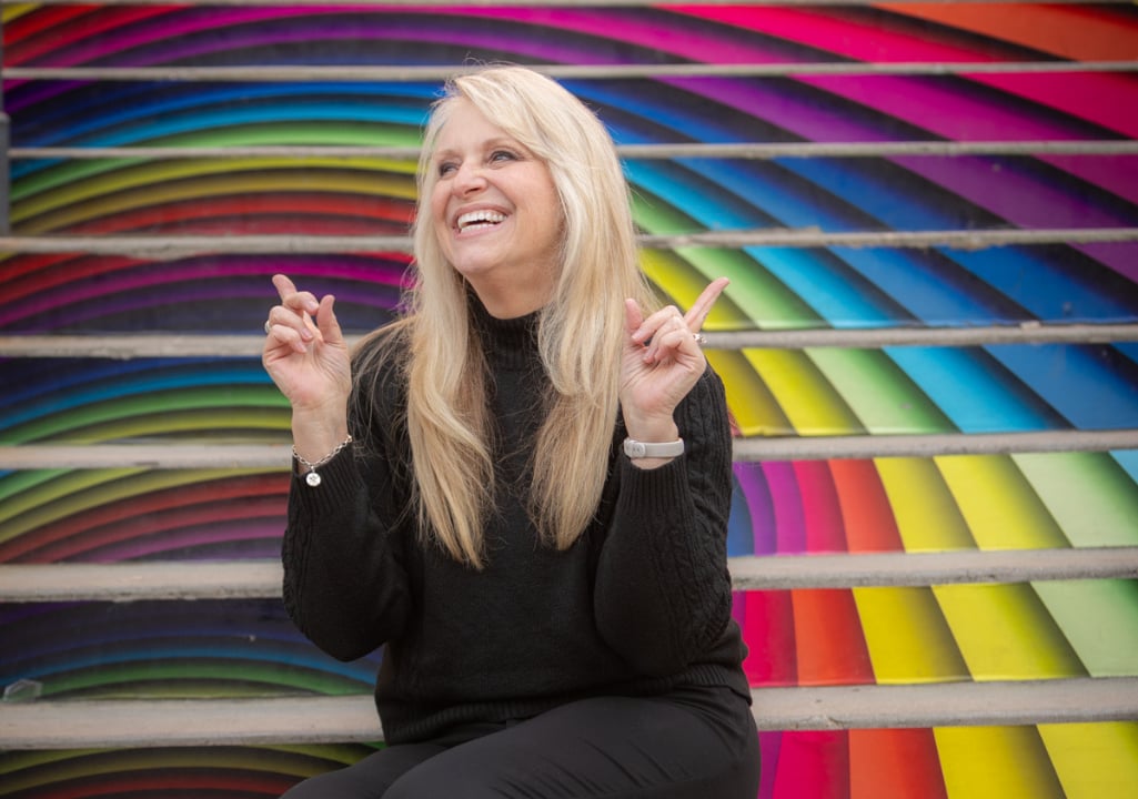 Sharla Jolley Ellis sitting on colorful rainbow-painted steps, smiling and pointing upward — a bright, joyful image representing positivity and optimism.