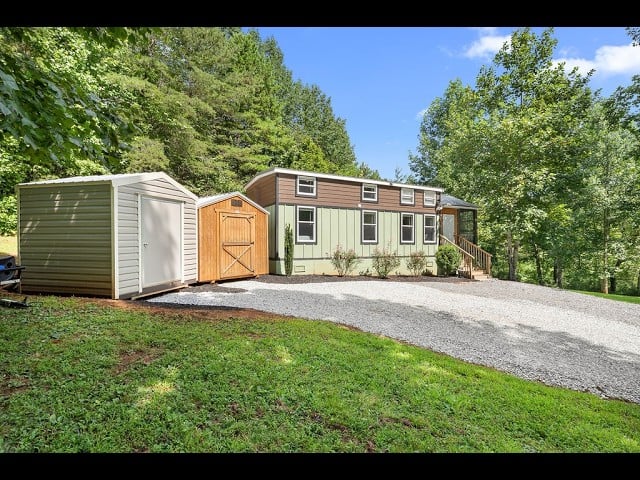 Manufactured home with two storage sheds beside a gravel driveway, surrounded by trees.