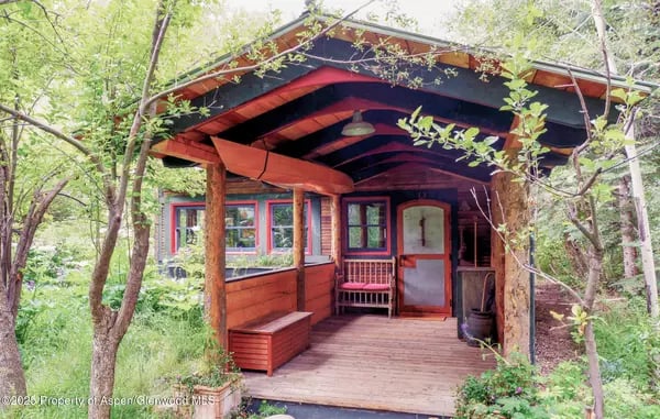 Rustic home with red trim and a covered wooden porch surrounded by leafy trees.