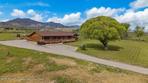 Wooden rural home beside a gravel drive and large shade tree, with mountains behind it.