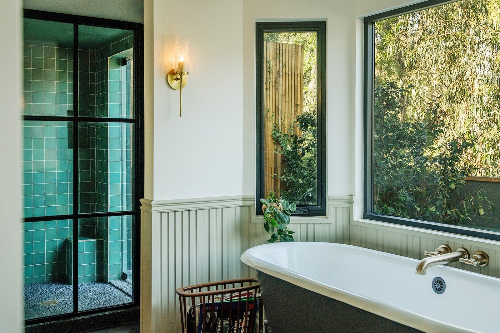 A bathroom with pale green beadboard wainscoting, a freestanding tub, brass fixtures and large windows looking onto trees.
