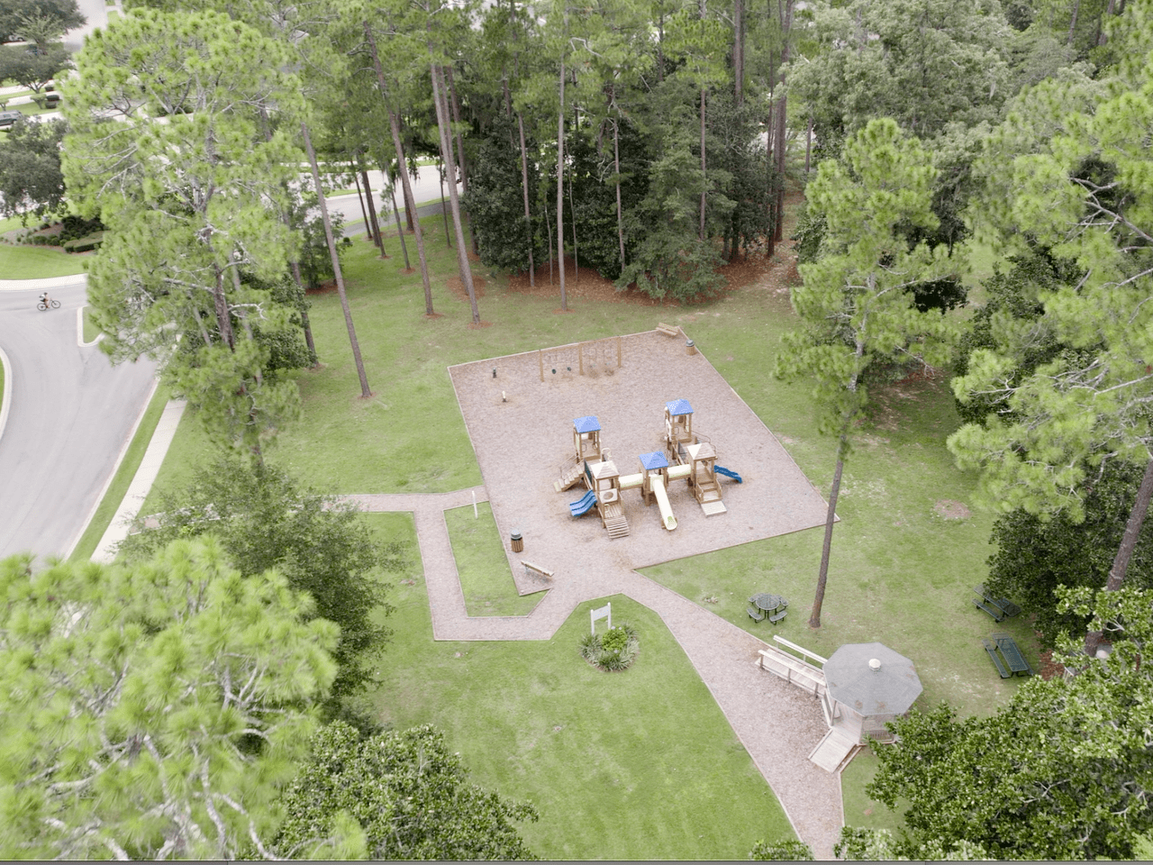 An aerial view of a playground area within a park. The playground features various equipment for children, such as slides and swings, surrounded by trees and green space. Paths are visible, leading to and from the playground.